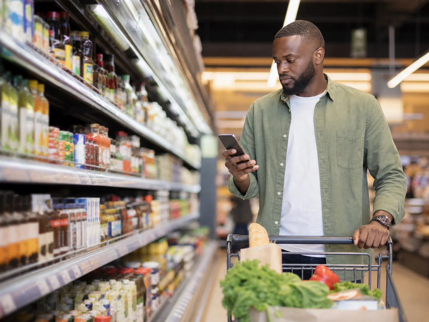 A shopper checks a phone beside a cart in a grocery aisle.